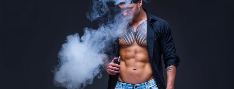 Vaper. The man dressed blue jeans, black shirt and black baseball cap with tattoos smoke an electronic cigarette on the dark background.