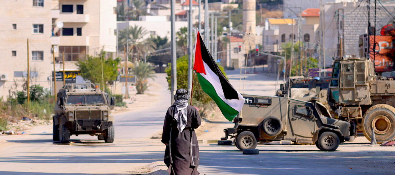 Un hombre lleva una bandera palestina mientras vehículos militares israelíes avanzan por una calle durante una redada llevada a cabo por fuerzas israelíes en la ciudad cisjordana de Tulkarem. Según el Ministerio de Salud palestino en Cisjordania, al menos un palestino murió y otros cinco resultaron heridos durante la redada en Tulkarem y su campo de refugiados.EFE/ Alaa Badarneh