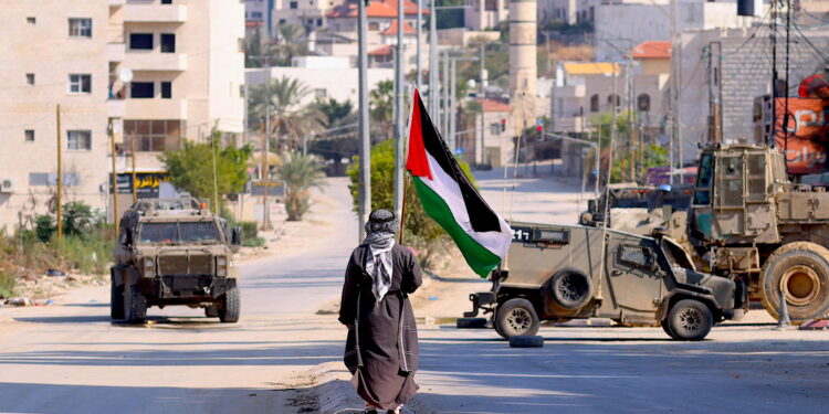 Un hombre lleva una bandera palestina mientras vehículos militares israelíes avanzan por una calle durante una redada llevada a cabo por fuerzas israelíes en la ciudad cisjordana de Tulkarem. Según el Ministerio de Salud palestino en Cisjordania, al menos un palestino murió y otros cinco resultaron heridos durante la redada en Tulkarem y su campo de refugiados.EFE/ Alaa Badarneh