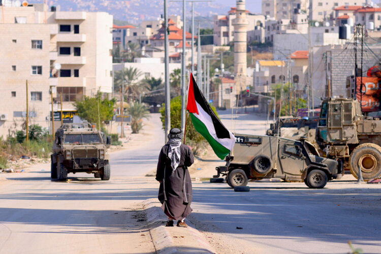 Un hombre lleva una bandera palestina mientras vehículos militares israelíes avanzan por una calle durante una redada llevada a cabo por fuerzas israelíes en la ciudad cisjordana de Tulkarem. Según el Ministerio de Salud palestino en Cisjordania, al menos un palestino murió y otros cinco resultaron heridos durante la redada en Tulkarem y su campo de refugiados.EFE/ Alaa Badarneh